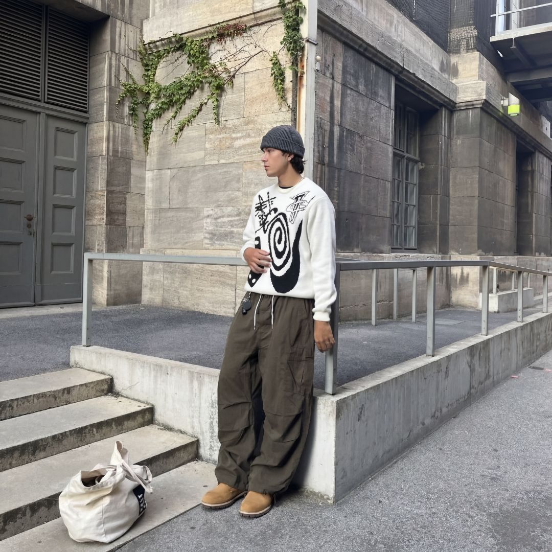 Mann lehnt an Geländer in urbanem Hintergund mit weißem Crewneck, grüner Hose und Timberland