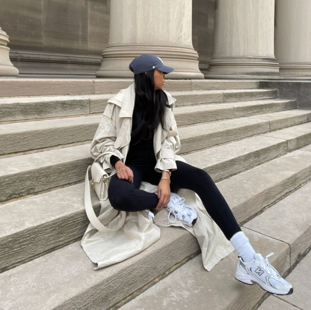 Frau sitzend auf einer Treppe mit weißen New Balance Sneaker und schwarzer Hose mit Beige Mantel
