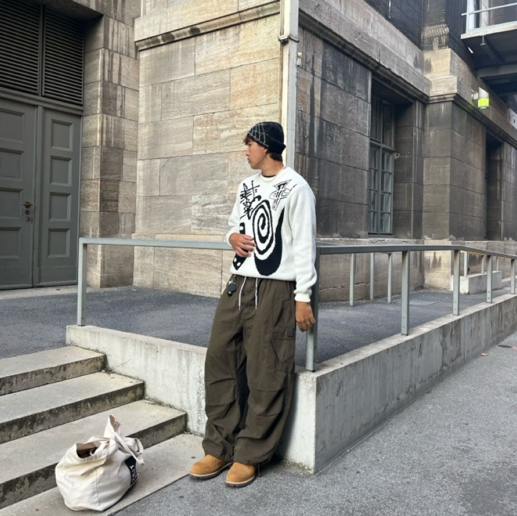 Mann lehnt an Geländer in urbanem Hintergund mit weißem Crewneck, grüner Hose und Timberland