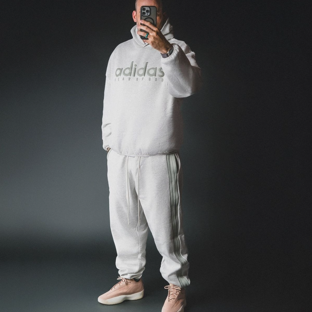 Mann vor schwarzem Hintergund mit Adidas x Fear Of God Schuhen, Jogginghose und Pullover in grau