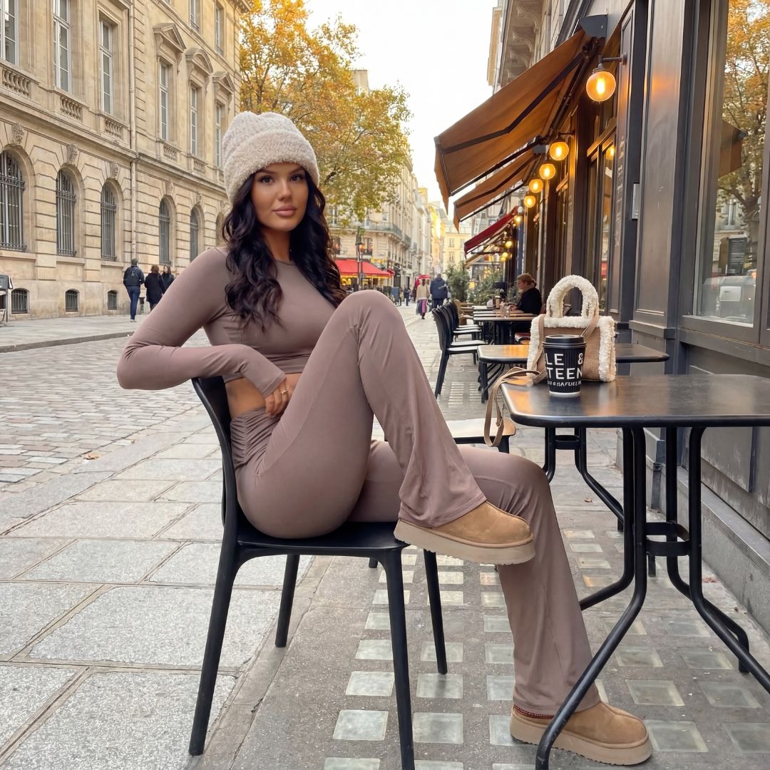 Frau sitzt auf Stuhl und trinkt einen Kaffee. Sie trägt ein braunes Outfit mit braunen Stiefeln und einer Mütze im Herbst.