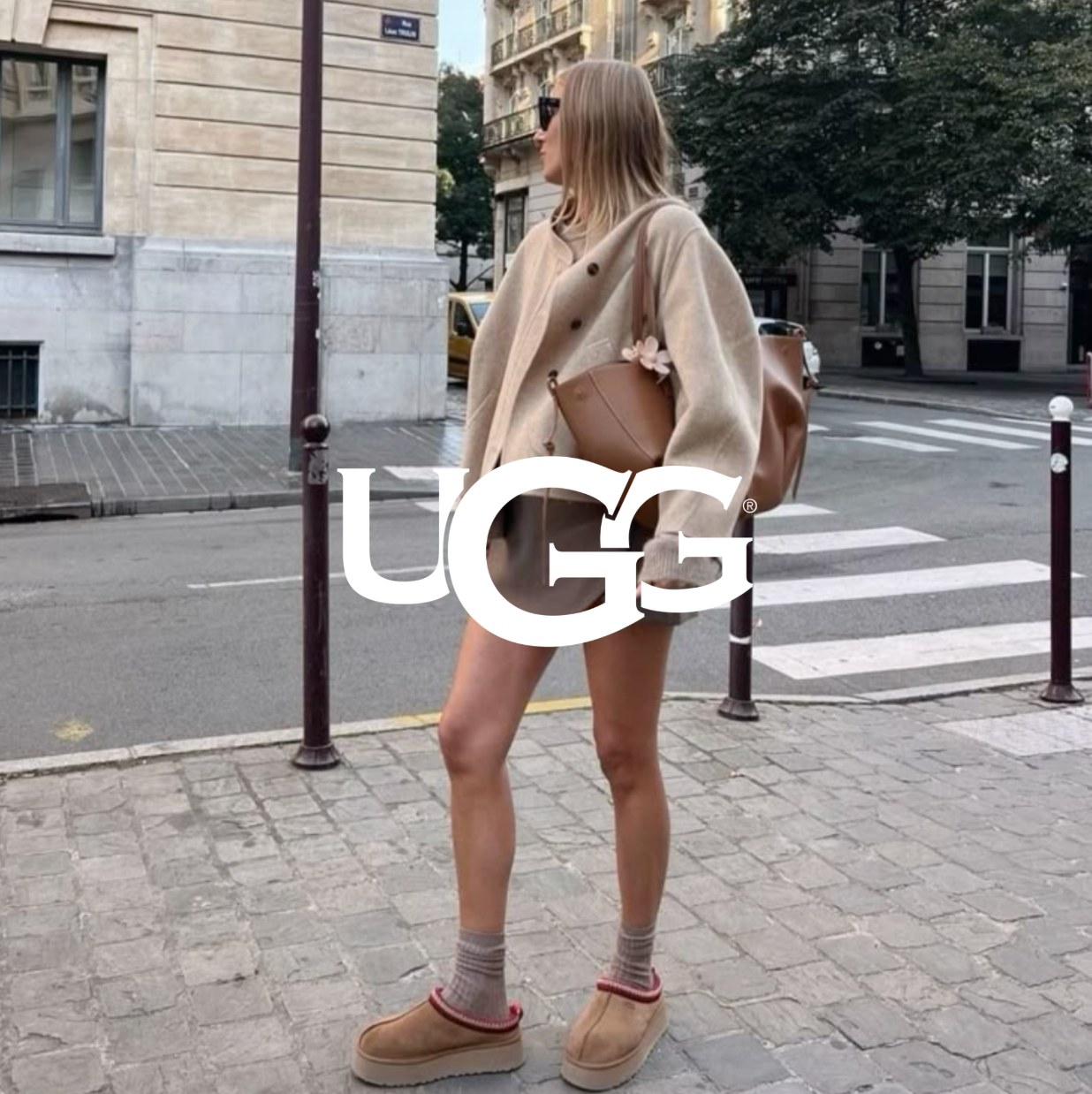 Person trägt UGG-Boots und einen beigen Cozy-Look beim Gehen durch eine urbane Straße; UGG-Logo im Vordergrund.