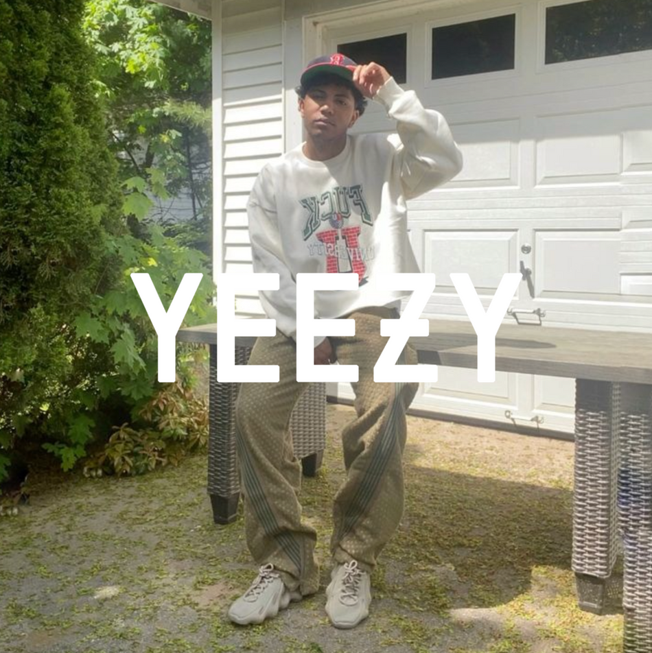 Alt-Text: Person in lässigem Streetwear-Outfit trägt YEEZY-Sneaker und posiert im Garten vor einer Garage; großes „YEEZY“-Logo im Bild.