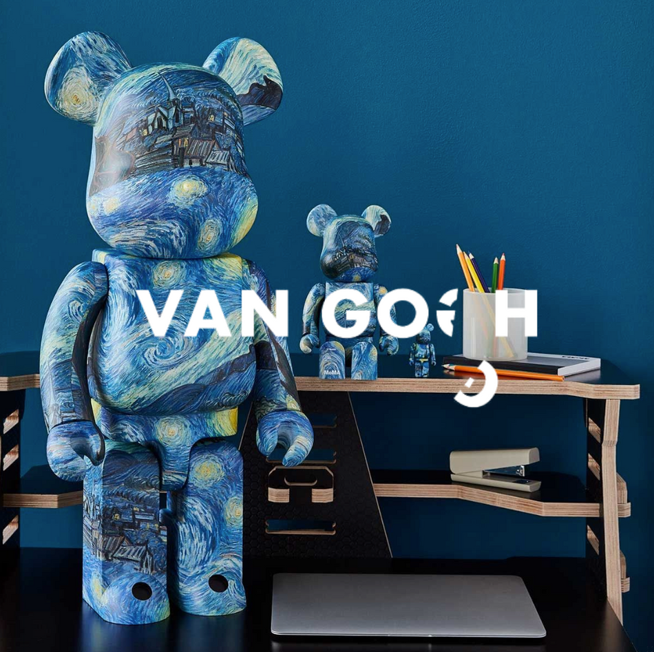 Bearbrick-Figur im Van-Gogh-Design mit „Sternennacht“-Motiv steht vor dunkelblauer Wand auf einem Schreibtisch; kleinere Figur im Hintergrund.