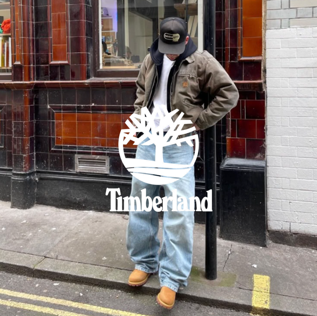 Timberland Logo, Mann steht mit blauer Jeans, braunen Boots und grüner Jacke vor einem Haus