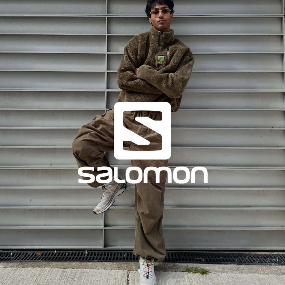 Salomon Logo, Mann vor einer Garage in grünem Streetwear Outfit mit Salomon Outdoor Schuhen
