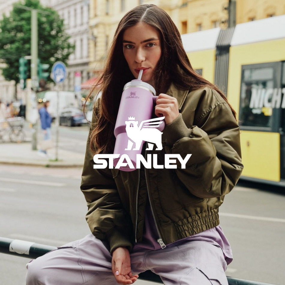 Stanley logo, Frau trinkend aus lila Stanley Becher sitzt auf geländer mit einer U-Bahn im Hintergrund