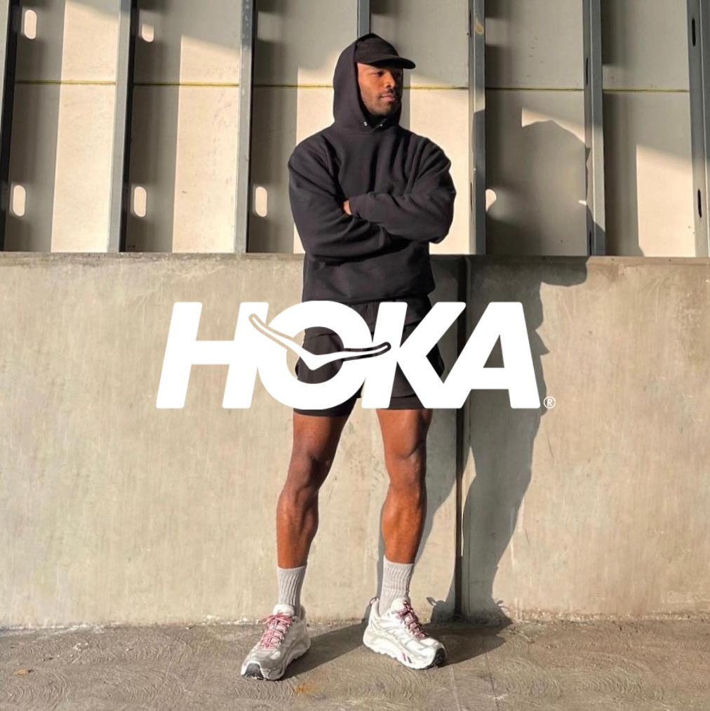 Hoka Logo mit einem Jogger mit Running Sneakern und schwarzem Sportoutfit stehend vor einem Gebäude