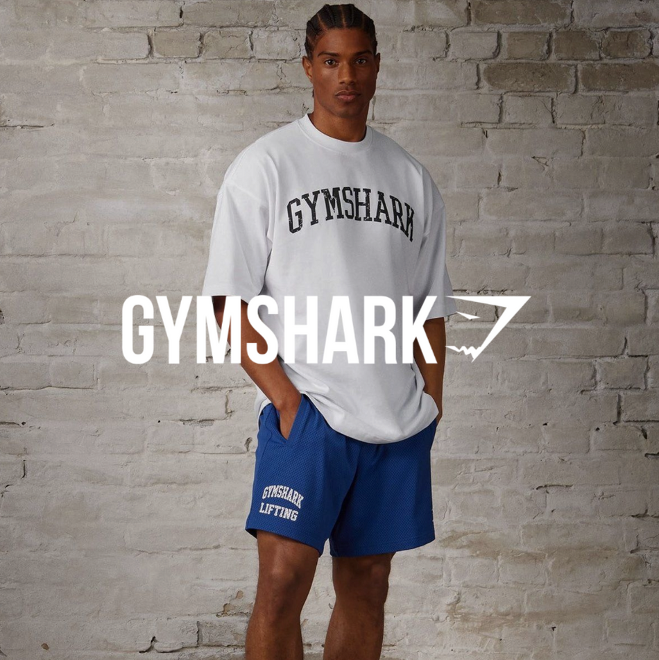 Gymshark Logo, Model mit blauer Sweatshorts und weißem T Shirt vor einer Steinwand