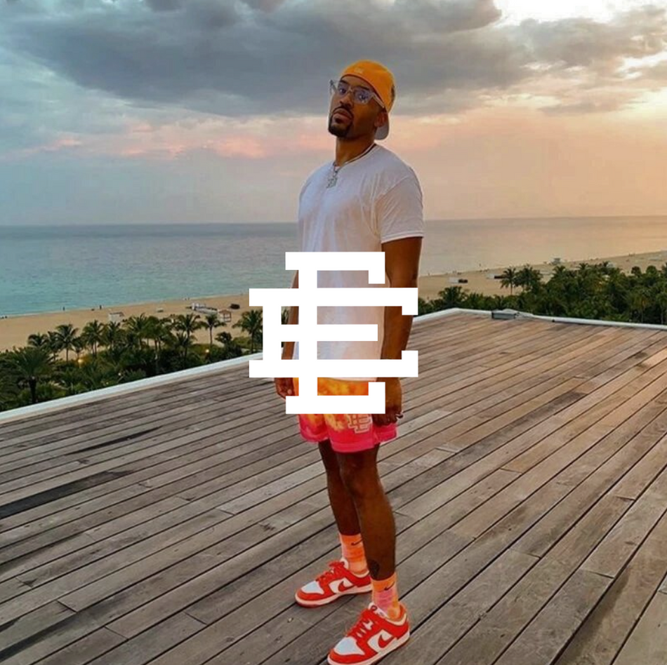 Eric Emanuel logo mit einem Mann stehend auf einer Terasse mit Strand im Ausblick. Er trägt rosa weiße SB Dunks und eine rosa eric emanuel shorts