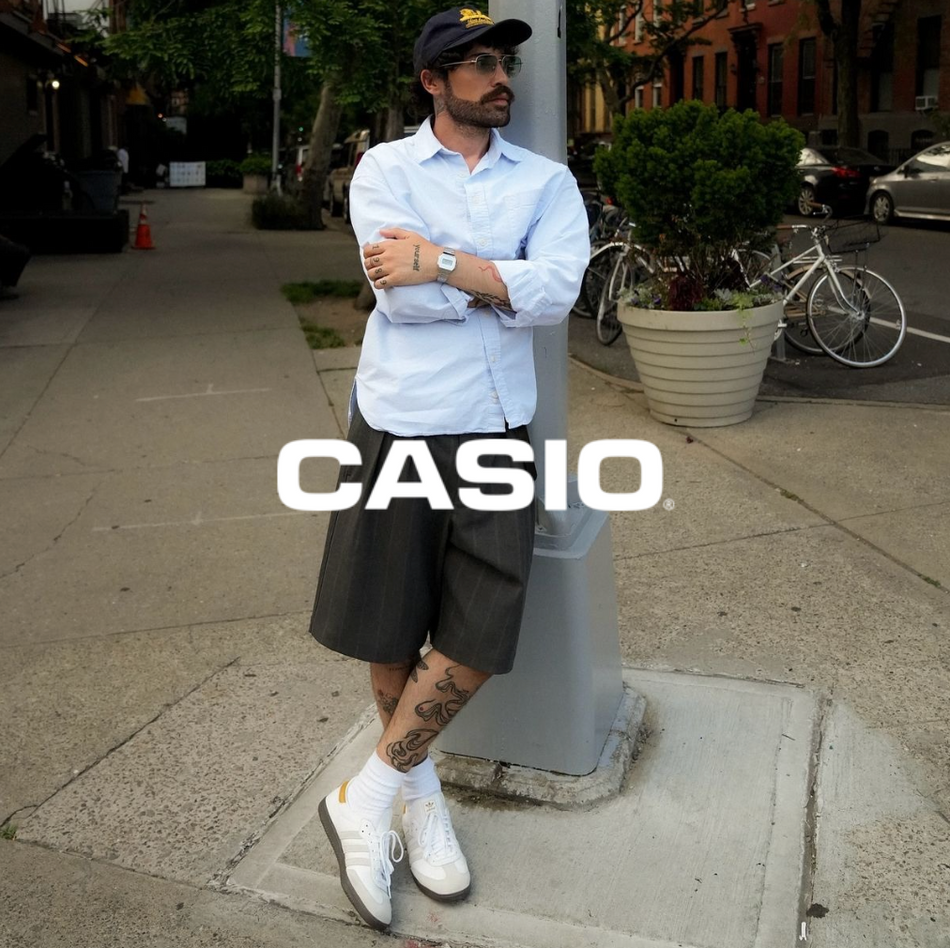 Casio Logo mit einem Mann in blauem Hemd mit schwarzer Shorts und Adidas Sneakern in weiß der eine Casio uhr trägt lehnend an einer Laterne