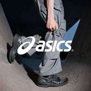 Asics Logo, dunkle Szene, mann mit dunklen Asics Sneakern steht vor beton Hintergund mit Cargopants in grau