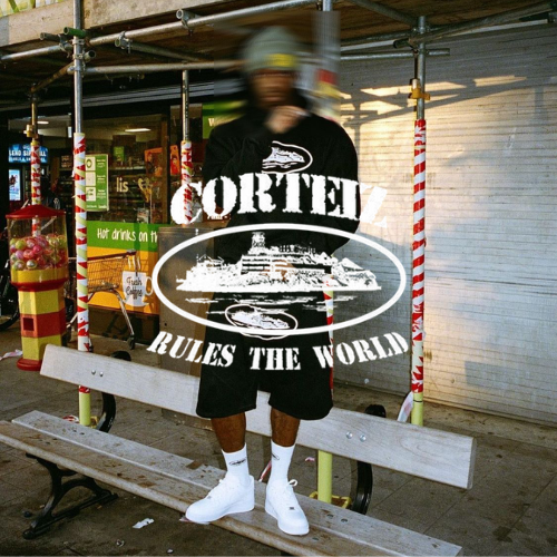 Corteize Logo, Mann in schwarzem Outfit mit weißen Nike Air Force 1 stehend auf einer Bank mit verschwommenen Look
