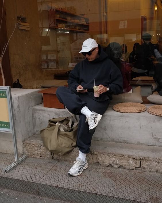 Mann vor Internetcafe mit weißen Asics Gel-1130 und schwarzem Outfit mit Handy in der Hand