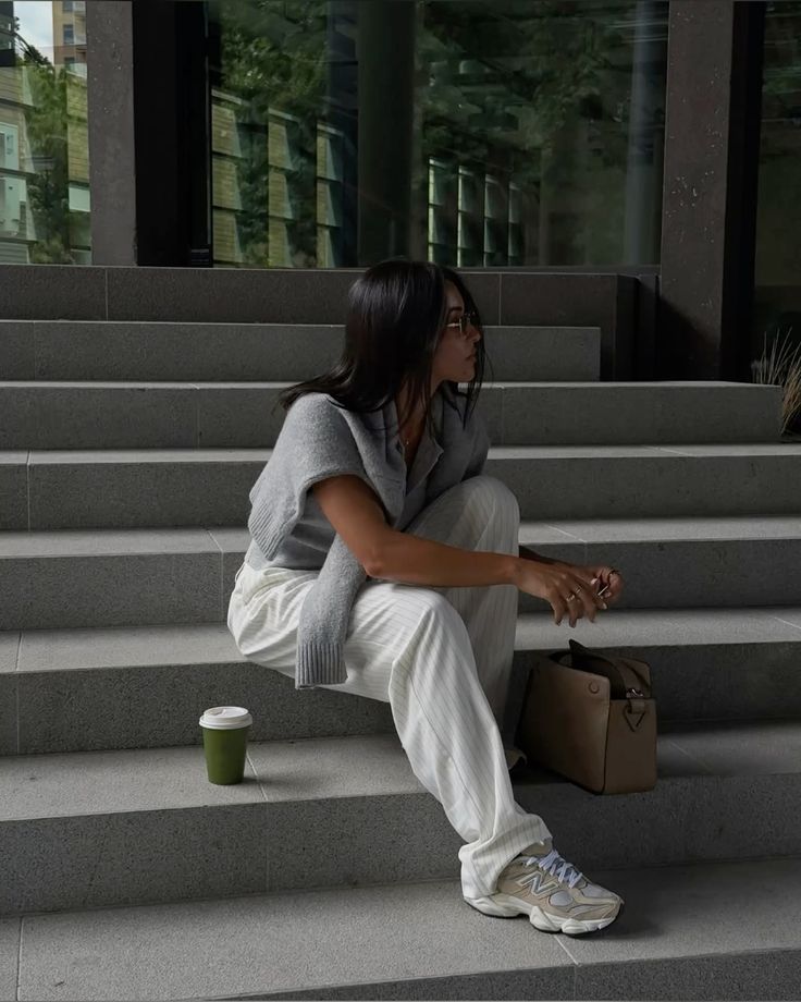 Frau mit grauem Pullover, weißer Hose und new Balance 9060 auf einer grauen treppe sitzend