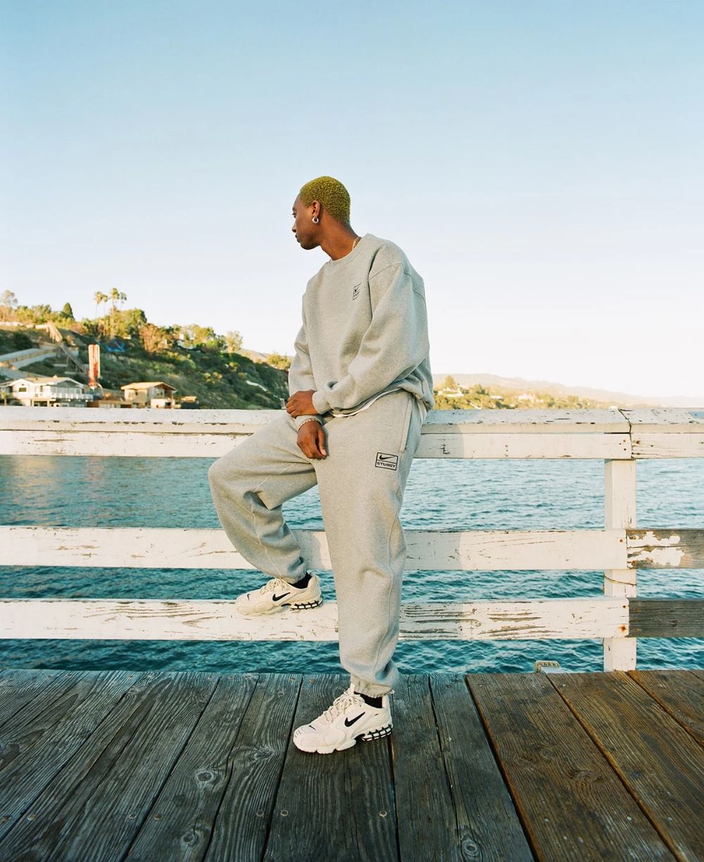 Mann auf Strandpromenade mit weißen Nike Sneakern, grauer Stussy Nike Jogginghose und passendem grauen Stussy nike Hoodie