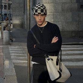 Model mit karierter Mütze und schwarzem Sweater und dunkler Jeans. Beige Tasche umgehängt und arme verschrenkt