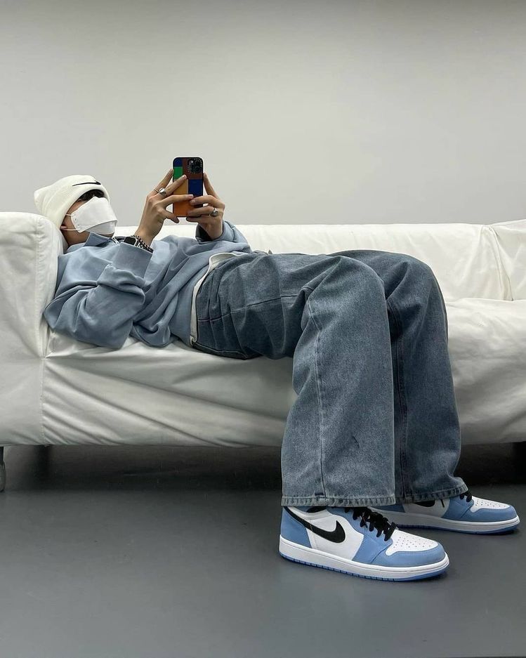 Mann liegend auf weißer Couch mit Jeans und Air Jordan 1 in blau sowie einem Sweater in blau schauend auf sein Handy